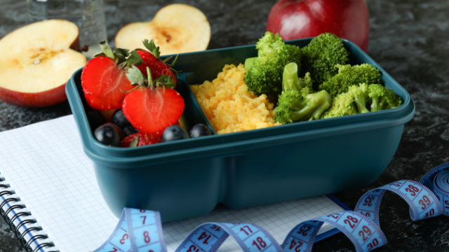 Zdjęcie przedstawia zdrowy posiłek w niebieskim pojemniku na lunch umieszczonym na białym zeszycie. Posiłek składa się z porcji kaszy jaglanej, świeżych brokułów i mieszanki owoców, w tym truskawek i jagód. Obok pojemnika znajduje się niebieska miarka krawiecka, sugerująca kontrolę porcji lub związek posiłku z dietą lub odchudzaniem. W tle widać przekrojone jabłko, co dodatkowo podkreśla zdrowy i zbilansowany charakter posiłku.