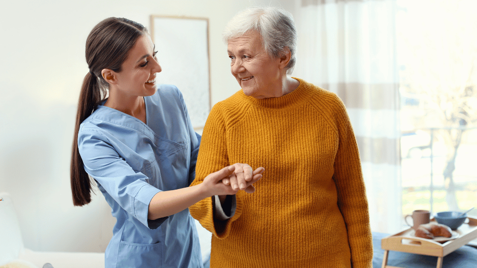 Młoda kobieta w niebieskim uniformie medycznym trzyma za rękę starszą panią w żółtym swetrze, obie stoją i uśmiechają się do siebie w jasnym, domowym otoczeniu.