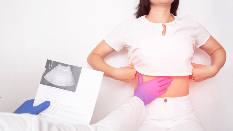 Kobieta podnosząca koszulkę, aby odsłonić brzuch, podczas gdy lekarz w niebieskich rękawiczkach dotyka jej boku, gdzie na dokumentach widoczne są wyniki badań ultrasonograficznych wątroby