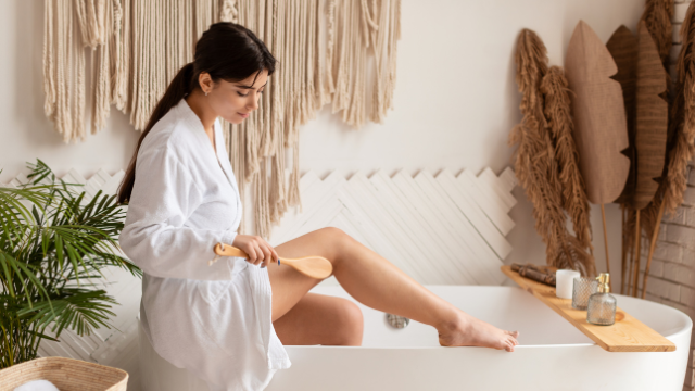 Kobieta w białym szlafroku wykonująca masaż antycellulitowy uda drewnianą szczotką w jasnej łazience, z elementami dekoracji w naturalnym stylu.