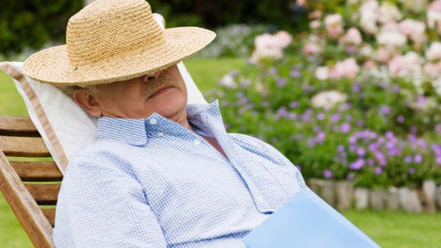 Starszy mężczyzna drzemie na drewnianym leżaku na zewnątrz w ogrodzie, z głową przykrytą słomianym kapeluszem, nosząc błękitną koszulę.