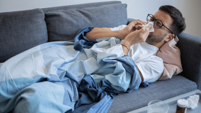 Mężczyzna z objawami przeziębienia lub grypy leży na kanapie pod kocem, dmucha nos w chusteczkę, a obok niego na kanapie widoczne są rozrzucone chusteczki i leki.