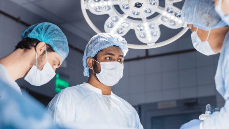 Chirurdzy w niebieskich strojach operacyjnych pracują w sali operacyjnej z widocznym oświetleniem operacyjnym.