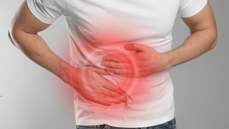 Mężczyzna w białej koszulce trzyma się za brzuch w okolicy żołądka, na którym nałożono czerwoną grafikę sugerującą ból lub dyskomfort w tej okolicy.