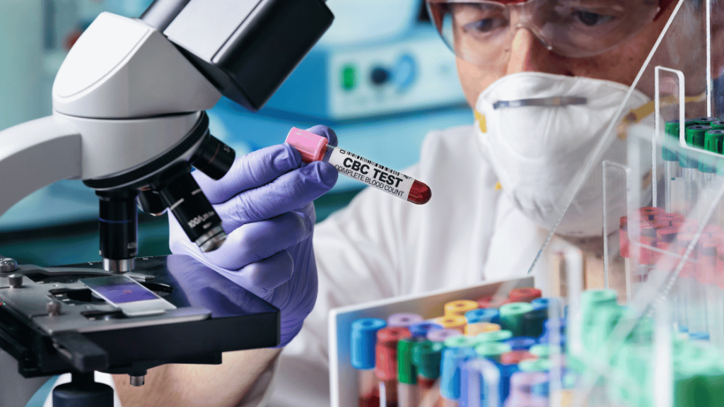 Naukowiec w laboratorium, noszący maseczkę ochronną i rękawiczki, trzyma probówkę z próbką krwi oznaczoną jako "CBC TEST" (morfologia krwi). W tle widoczny jest mikroskop oraz stojak z różnokolorowymi probówkami.