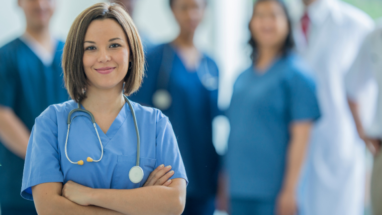 Młoda lekarka w niebieskim mundurku medycznym z założonymi rękami, w tle zespół medyczny.